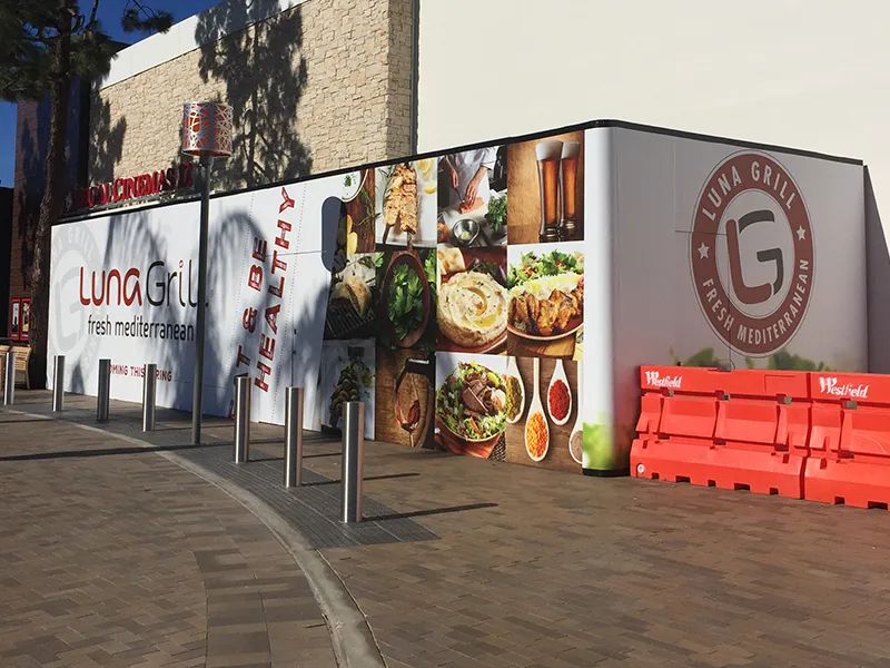 A construction hoarding wrapping a corner site, carrying the wordmark, a campaign line, and a grid of food photography