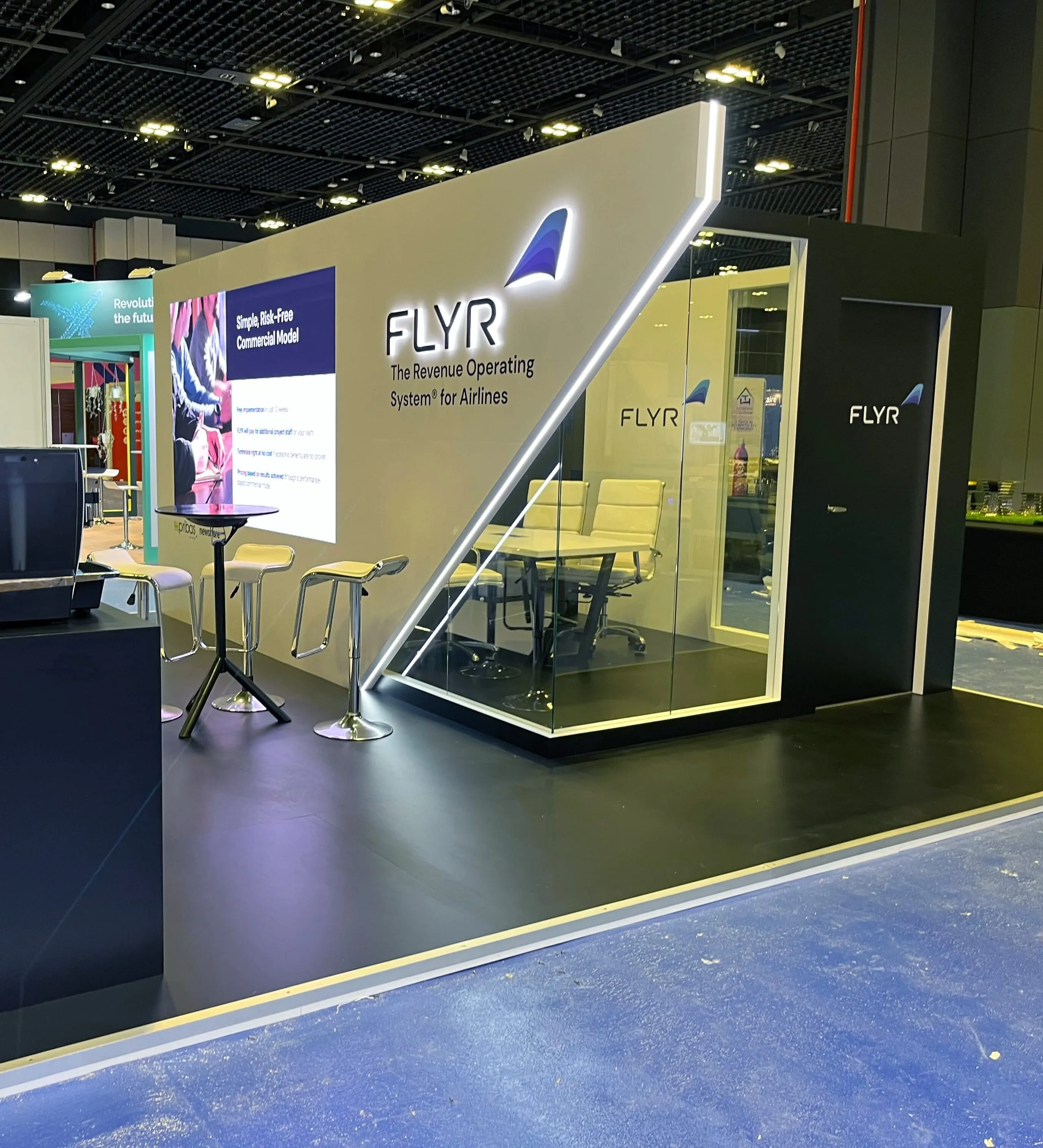The 2023 variation of the booth: the same wall system and logo treatment, with an enclosed glass meeting room built into the footprint.